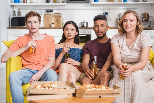 multicultural friends with bottles of beer watching tv near pizza during party - 写真・画像