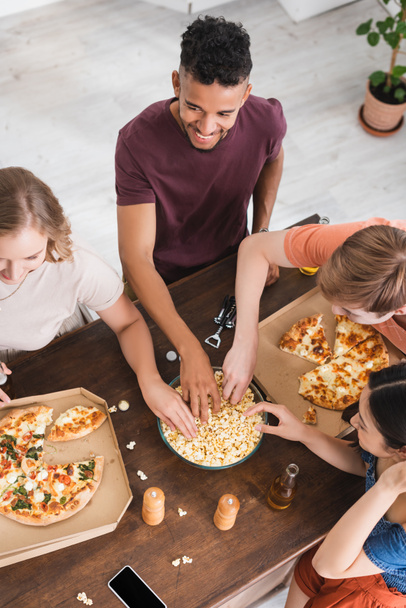 overhead view of multicultural friends taking popcorn near pizza on table - Foto, Imagen
