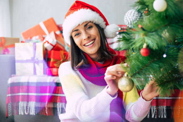 Close up photo of a beautiful happy young woman in a sweater and santa hat is decorating a christmas tree at home on gift boxes background. Winter holidays - Fotó, kép