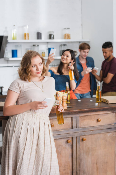 enfoque selectivo de la mujer joven sosteniendo cerveza y teléfono inteligente cerca de amigos multiétnicos en el fondo - Foto, Imagen