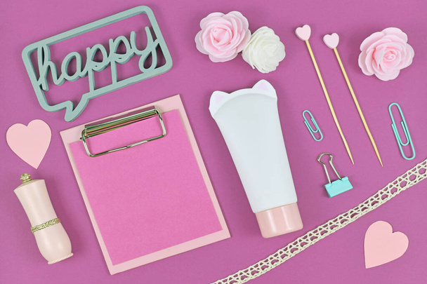 Flat lay with blank clipboard and various cute items like rose flowers, cream tube in shape of cat, paper hearts andwooden word 'Happy' on purple background - Foto, Imagen
