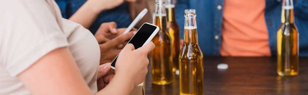 częściowy widok wieloetnicznych przyjaciół czatujących na smartfonach przy piwie podczas imprezy, uprawa pozioma - Zdjęcie, obraz