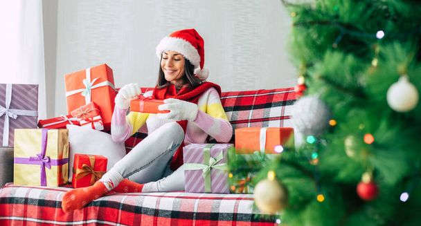 Celebration of the winter holidays. Beautiful happy brunette woman in santa hat and sweater is having fun with many gift boxes on the couch at home. Merry Christmas and happy new year - Photo, image