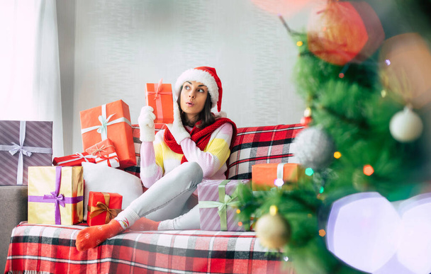 Celebration of the winter holidays. Beautiful happy brunette woman in santa hat and sweater is having fun with many gift boxes on the couch at home. Merry Christmas and happy new year - Foto, immagini