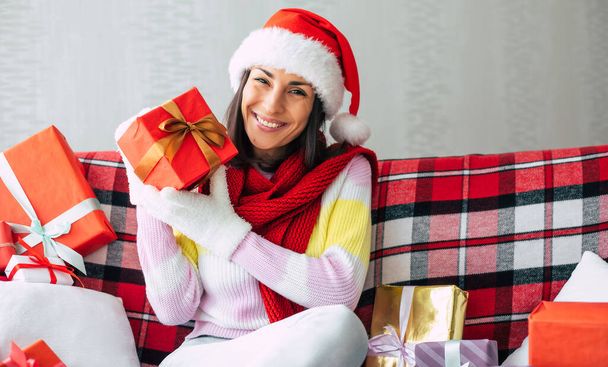 Celebration of the winter holidays. Beautiful happy brunette woman in santa hat and sweater is having fun with many gift boxes on the couch at home. Merry Christmas and happy new year - Photo, Image