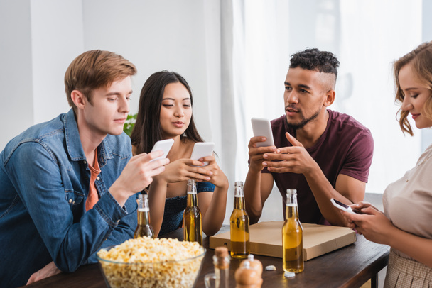 selective focus of multicultural friends chatting on smartphones near beer and popcorn during party - Foto, imagen
