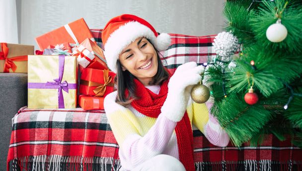 Close up photo of a beautiful happy young woman in a sweater and santa hat is decorating a christmas tree at home on gift boxes background. Winter holidays - Fotoğraf, Görsel