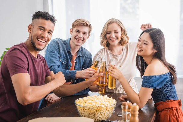 joyful multicultural friends looking at camera while clinking bottles of beer - Photo, image