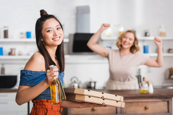 enfoque selectivo de la mujer asiática sosteniendo cajas de pizza y cerveza cerca de amigo en el fondo - Foto, Imagen
