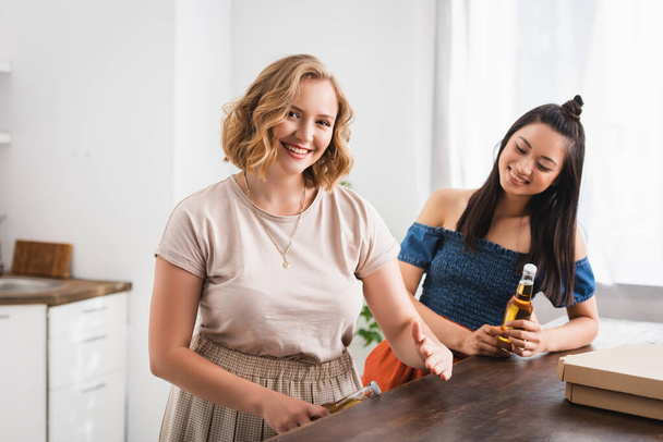 joyful woman opening bottle of beer near asian friend during party - Photo, image