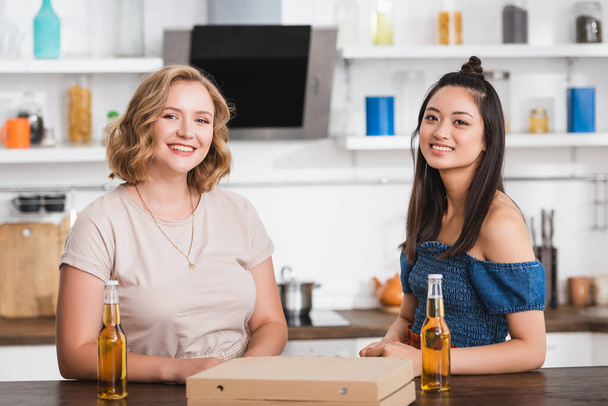 junge multikulturelle Frauen blicken in die Kamera neben Bier- und Pizzaschachtel auf dem Tisch - Foto, Bild