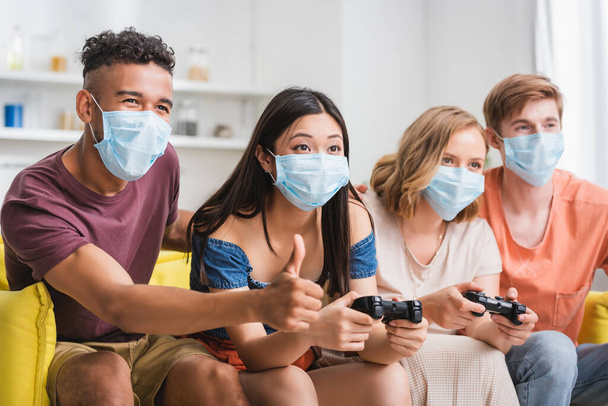 KYIV, UKRAINE - JULY 28, 2020: african american man showing thumb up near multicultural friends playing video game in medical masks - Фото, зображення
