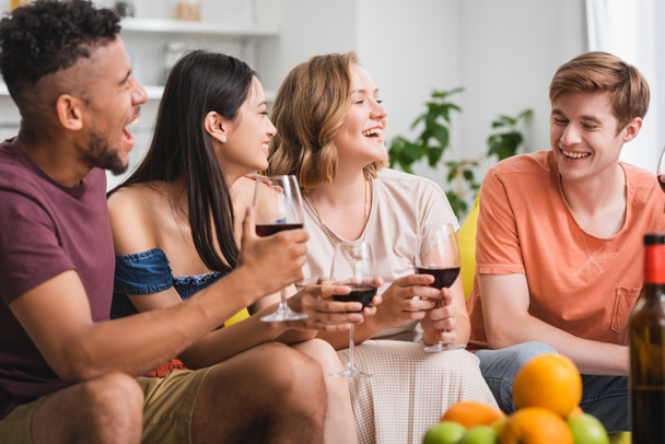 excited multicultural friends talking while holding glasses of red wine in kitchen - Photo, Image