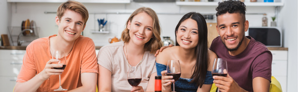 horizontal image of multiethnic friends looking at camera while holding glasses of red wine in kitchen - Photo, Image