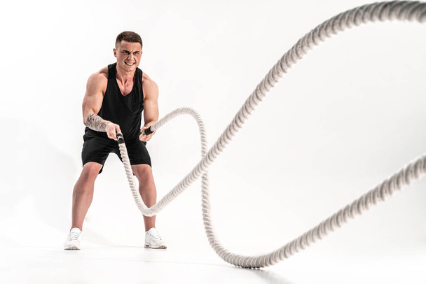 Attractive muscular man working out with heavy ropes. Photo of handsome man in sportswear isolated on white background - Photo, Image