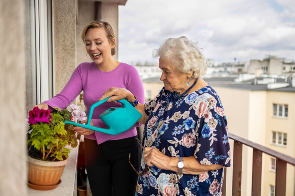 Oudere vrouw en haar volwassen kleindochter besproeien planten op het balkon - Foto, afbeelding