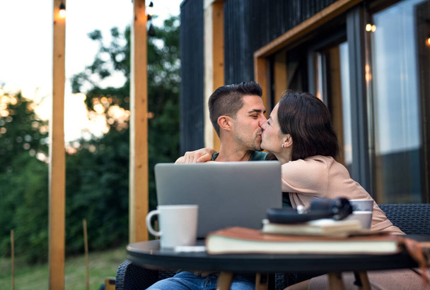 Young couple with laptop kissing outdoors, weekend away in container house in countryside. - Foto, afbeelding