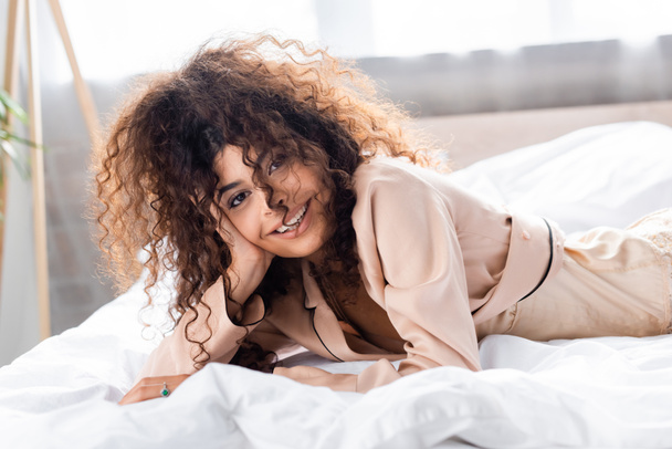 joyful and curly woman looking at camera while lying on bed - Фото, изображение