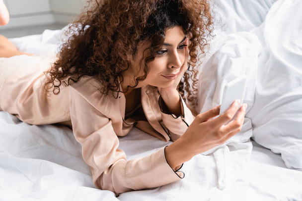 молодая кудрявая женщина лежит на кровати и использует смартфон в спальне - Фото, изображение