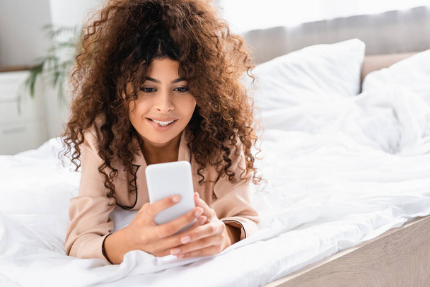 young curly woman biting lip and using smartphone in bedroom - Foto, imagen