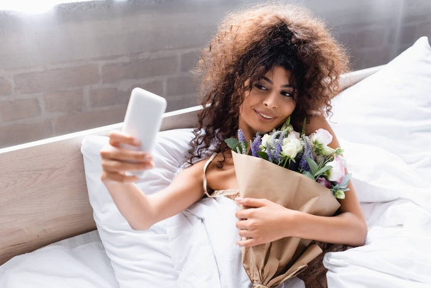 enfoque selectivo de la mujer complacida tomando selfie con flores en el dormitorio - Foto, Imagen
