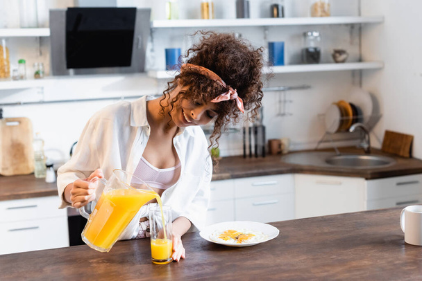 σγουρή και χαρούμενη γυναίκα που κρατάει κανάτα και ρίχνει φρέσκο χυμό πορτοκαλιού κοντά σε ποτήρι και πιάτο με τηγανητά αυγά - Φωτογραφία, εικόνα