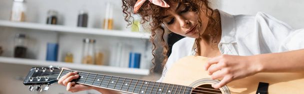 panoramic crop of curly woman playing acoustic guitar at home - Fotografie, Obrázek