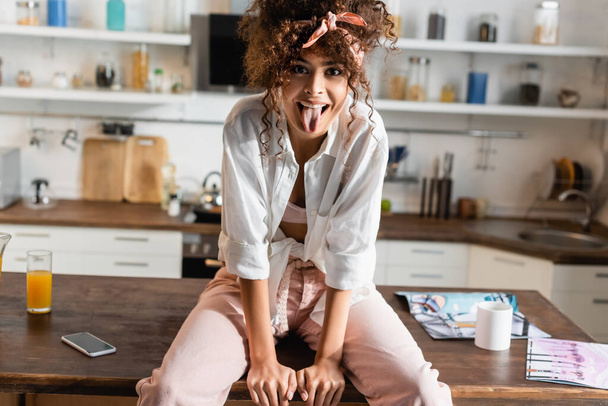 curly woman sitting on table and sticking out tongue  - Fotó, kép