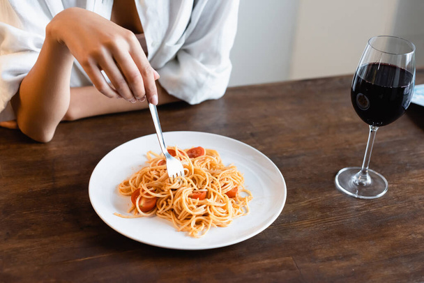 μερική άποψη της γυναίκας κρατώντας πιρούνι κοντά στο πιάτο με έτοιμα μακαρόνια και ένα ποτήρι κόκκινο κρασί - Φωτογραφία, εικόνα
