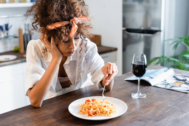 upset woman holding fork near plate with prepared spaghetti and glass of wine on table - Foto, afbeelding