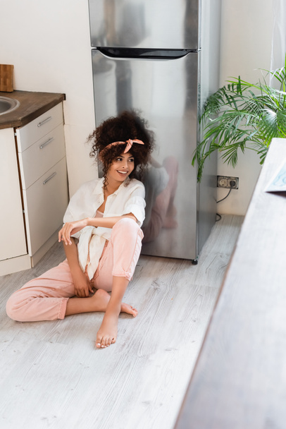animado e descalço mulher sentado no chão da cozinha perto de geladeira - Foto, Imagem