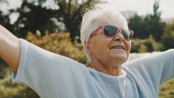 Portrait de femme âgée heureuse avec des lunettes de soleil roses avec les mains tendues à l'écoute de la musique à l'extérieur - Photo, image