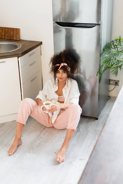 curly woman sitting on kitchen floor with cup of coffee - Photo, image
