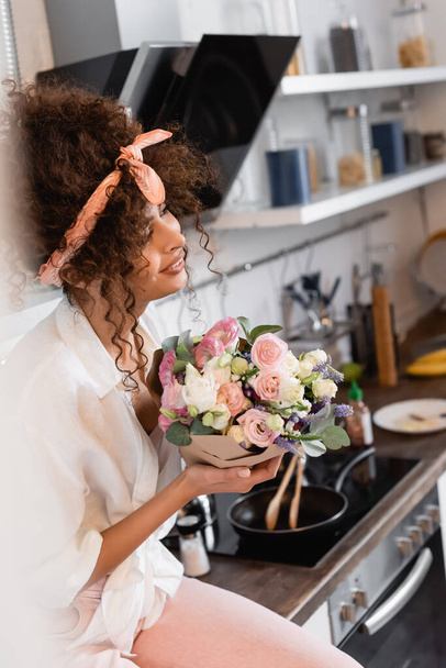 selective focus of curly young woman holding flowers in kitchen  - Fotó, kép