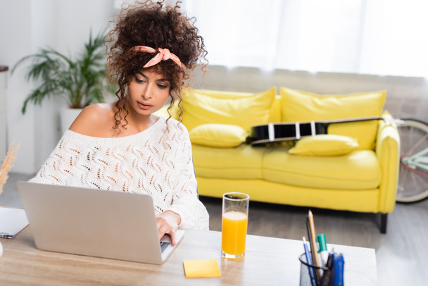 curly freelancer looking at laptop near glass of orange juice  - Photo, Image