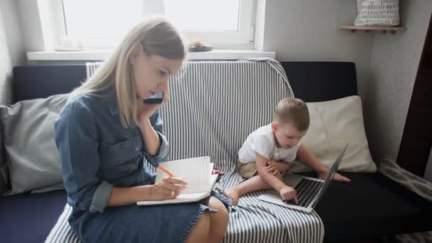 Druk bezig vrouw praten over de telefoon en het maken van notities, terwijl haar zoon speelt met notebook - Video
