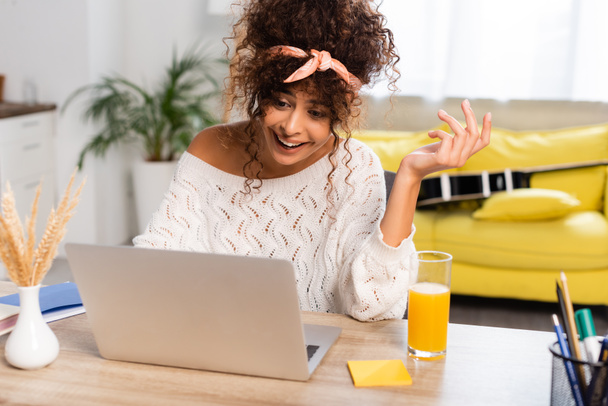 joyful woman using laptop near vase with fluffy branches and glass of orange juice - Photo, Image