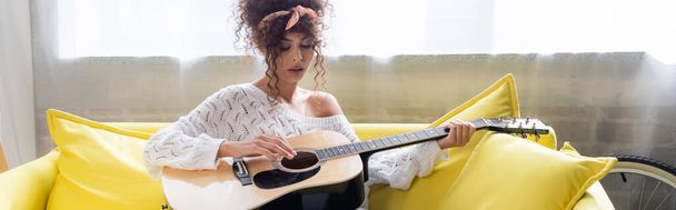 panoramic shot of curly woman playing acoustic guitar on sofa in living room  - Foto, Imagen