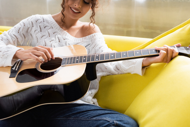 oříznutý pohled na mladou kudrnatou ženu hrající na akustickou kytaru na pohovce v obývacím pokoji  - Fotografie, Obrázek