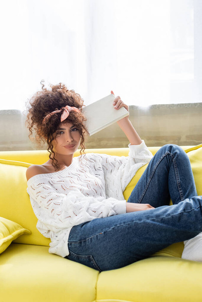 curly young woman holding book and looking at camera in living room  - 写真・画像