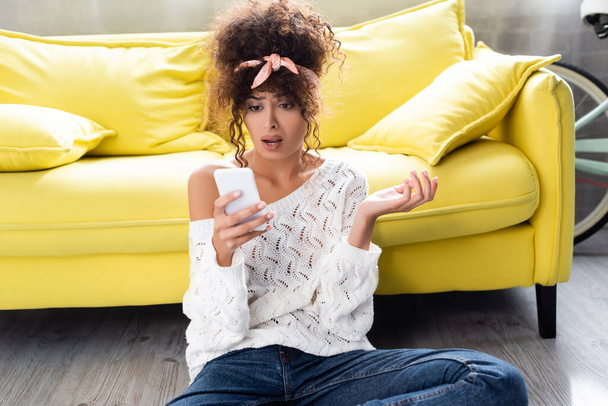 Unzufriedene Frau blickt auf Smartphone neben gelbem Sofa - Foto, Bild