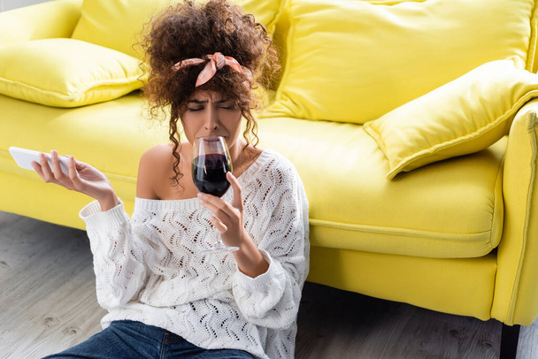 Verärgerte Frau hält Glas Wein und Smartphone in der Hand und weint im Wohnzimmer - Foto, Bild
