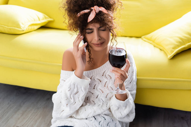 lockige Frau hält ein Glas Wein in der Hand und spricht auf dem Smartphone, während sie im Wohnzimmer weint - Foto, Bild