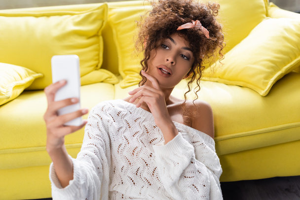 selective focus of curly woman taking selfie in living room  - 写真・画像