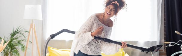 panoramic shot of curly woman having fun while holding vacuum cleaner and cleaning home - Foto, imagen