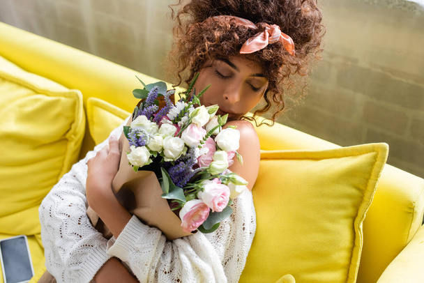 目を閉じて花束を持ち居間で花の匂いを嗅ぐ若い女性 - 写真・画像