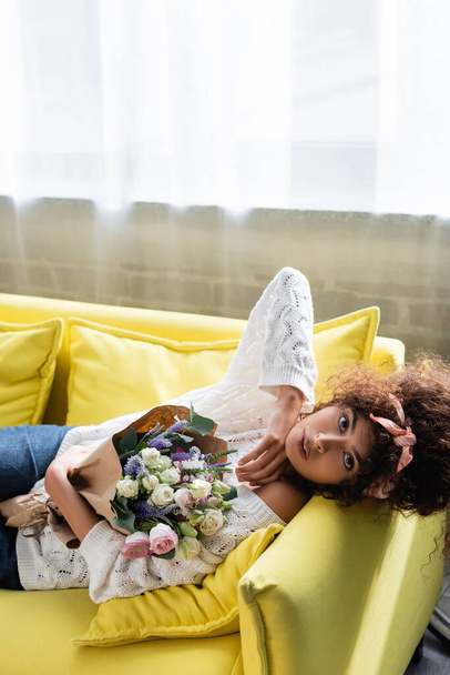young curly woman lying on yellow sofa with flowers  - Photo, Image