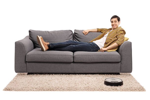 Young man sitting on a sofa and a robotic vacuum cleaner vacuuming the carpet isolated on white background - Foto, Imagem