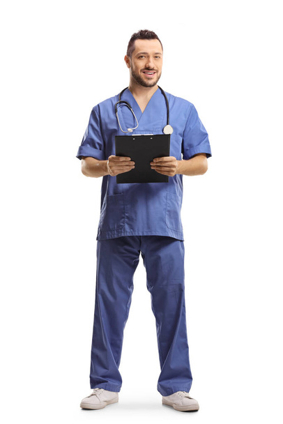 Full length portrait of a male doctor in a blue uniform holding a clipboard and smiling isolated on white background - Zdjęcie, obraz