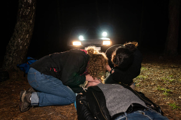 Two women are helping a man lying on the ground. Night scene in the headlights of a car.  Field First Aid Training - Foto, Imagen
